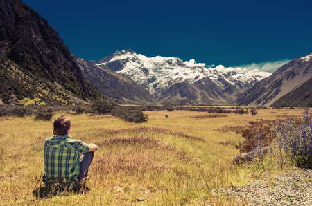 Rear view of a man sitting on grass and looking at mountains and glacierの写真素材