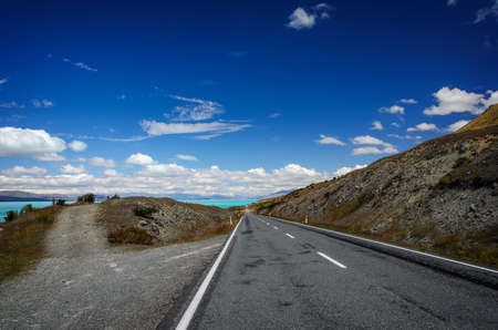 Straight empty road through fields to the Lake Tekapo and blue sky with white clouds. New Zealandの写真素材
