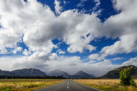 Straight empty road through fields to the mountain and deep blue sky with big white clouds.. New Zealandの写真素材