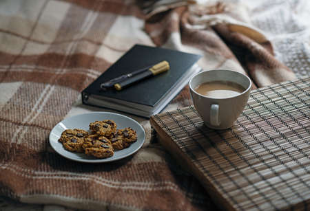 Scene with coffee on the table, oatmeal cookies with chocolate chips and a notebook with a pen and marker on the bed with plaidの写真素材