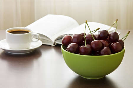 Bowl of sweet cherries, cup of coffee and book on table against windowの写真素材