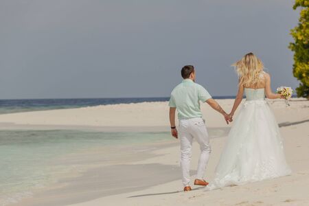 Young married couple walking along the ocean shore in Maldives. They gently holding each other's hands. The bride with flowers wearing a turquoise dress. Wedding and romantic honeymoon concept.の写真素材