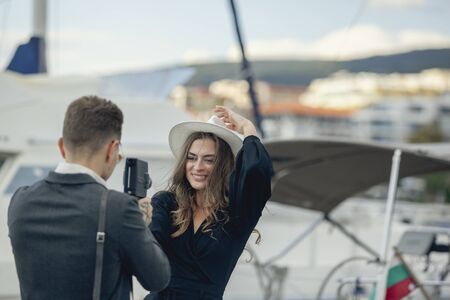 Young man taking pictures of his girlfriend on retro vintage camera. Attractive girl in hat smiling and posing for the best picture. Happy romantic married adult couple having fun, playing, fooling.の写真素材
