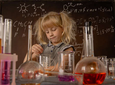 Curious little girl with test tubes and colorful substances makes tests at school laboratory. Small...