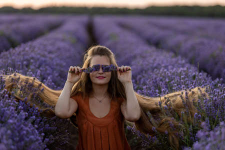 Smiling pretty romantic girl with long hair and in orange dress holds a bunch of lavender flowers and closes her eyes with them. Girl sits in a violet lavender field at sunset. Summer travel concept.の写真素材