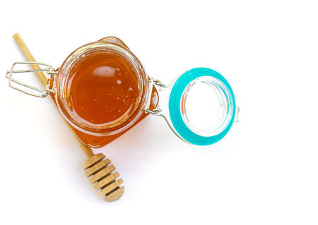 Opened glass jar of floral golden honey with lid wooden honey stick on white background
