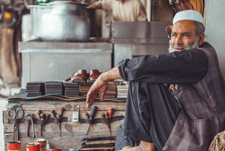 Poor old sad Pakistani Pathan shoe man cobbler on the local streets of Pakistan with his hand made leather shoes and repair tools in his street shopの写真素材