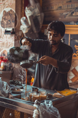 Pakistani man food vendor preparing and pouring smoking hot masala chai, the local tea, into cups...