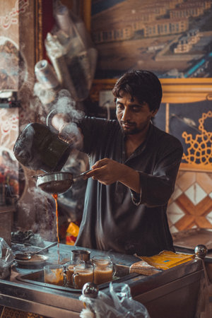 Pakistani man food vendor preparing and pouring smoking hot masala chai, the local tea, into cups from kettle in his roadside street food stallの写真素材