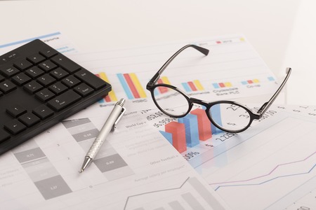 Financial printed paper charts, graphs on desk with pen, mobile, glasses and keyboardの写真素材