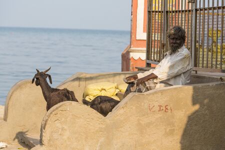 RAMESHWARAM, TAMIL NADU, INDIA - MARCH CIRCA, 2018. a goat walks by a homeless man sitting on the streets, near the gate and the beachのeditorial素材