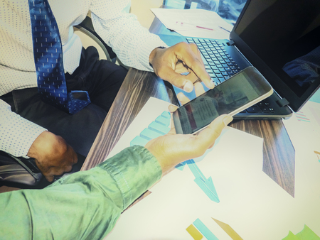 Two business young indian adults men with smartphone and laptop working and communicating while sitting at the office desk with graphs sheet, together with colleagues. Vintage treatment. Aerial viewの写真素材