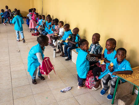 MBOUR, SENEGAL - JANUARY Circa, 2021. Little unidentified black cute kids children with uniforms are sitting on a bench in their primary school. Having breakfast during recreation.のeditorial素材