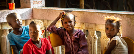 MBOUR, SENEGAL - DECEMBER, Circa, 2020. Unidentified group of cheerful children sitting on chairs outdoors, looking at teacher to learn song with musicans. Sunny day, selective focus.のeditorial素材