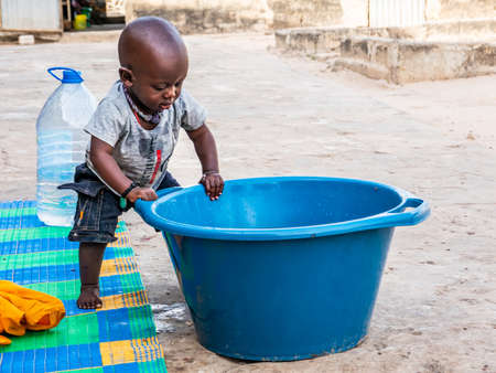 MBOUR, SENEGAL - DECEMBER Circa, 2020. African baby, less than one year, wanting bathing in a basin in a poor village senegalese house. Turning around big blue bowl to play with water.のeditorial素材