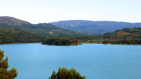 Large reservoir lake in the mountains, very blue water, pine trees on the shore, an island in the distance, mountains in the distance, beautiful landscape, Beniarres, Spainの写真素材