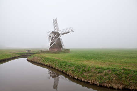 Dutch wooden windmill in fog by canalの写真素材