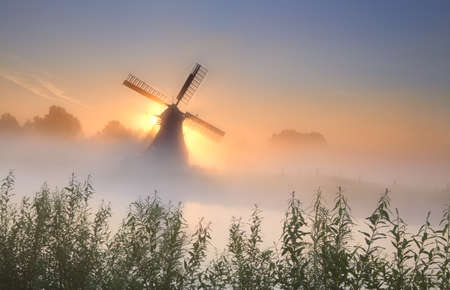 misty sunrisebehind the windmill near riverの写真素材