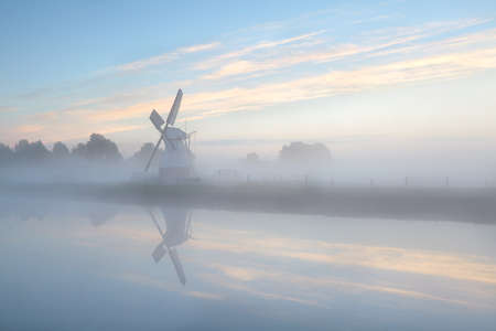 Dutch windmill in dense fog during sunrise by river, Hollandの写真素材