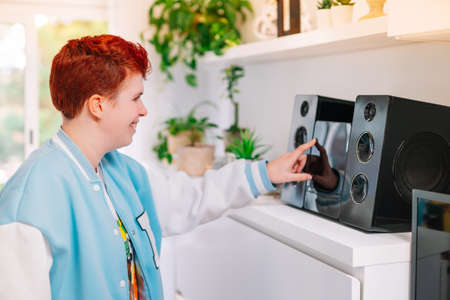 Modern, short-haired, red-haired, non-binary woman, turns on mini-system at home.の写真素材