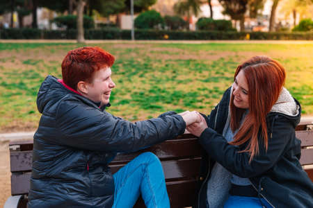 Couple of two lesbian women, smiling, sitting, holding hands on a bench in a public park.の写真素材