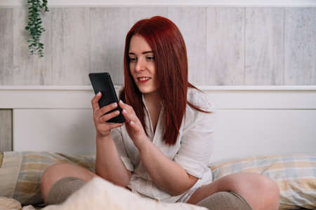 smiling young woman, using her cell phone, sitting on her bedroom bed.