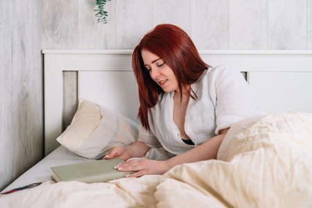 Sexy young woman lying on her bedroom bed, reading a book.