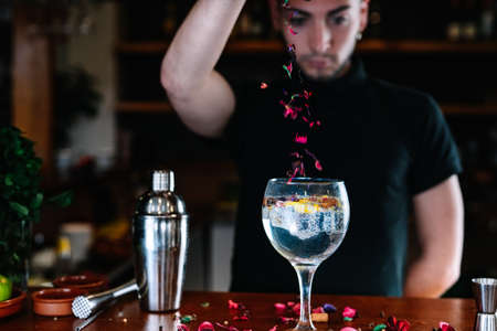Young bartenders preparing a mixed drink in a crystal glass. Depositing flowers of toppings.