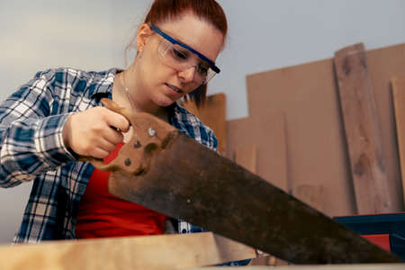 close-up of a young red-haired woman carpenter sawing a wooden board with a saw, in her small carpentry shop.の写真素材