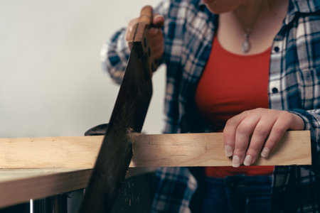 Close-up cropped shot of a young red-haired female carpenter, sawing a plank of wood in her small workshop.の写真素材
