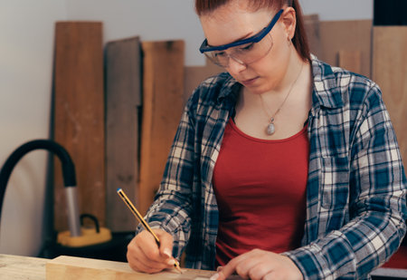 Young red haired carpenter woman, measuring a plank of wood in her small carpentry workshop.の写真素材