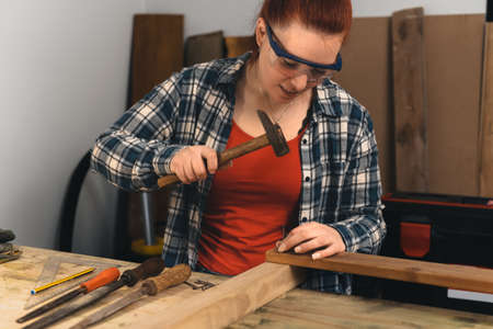 Young red-haired carpenter hammering a nail into a wooden board in her small carpentry workshop.の写真素材