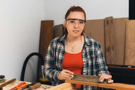 Young red-haired female carpenter using a precision level in her small carpentry workshop. Looking...