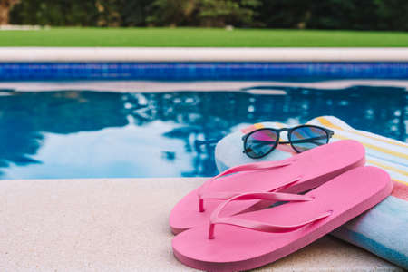 summery swimming objects in front of a swimming pool. beach towel, sunglasses, swimming flip-flops. lawn garden with swimming pool in the background.の写真素材