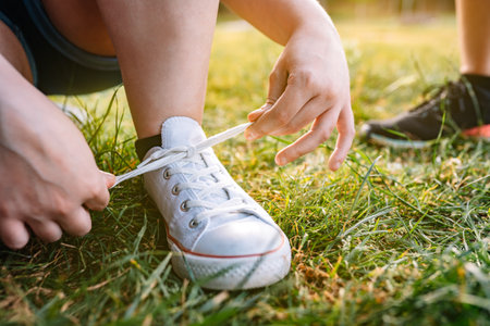 close-up of the hands of a young girl bending down, tying her white trainers in a public park in the...