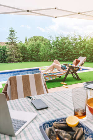 Scene of a table on the terrace of a house in summer with a woman lying by the pool in the...
