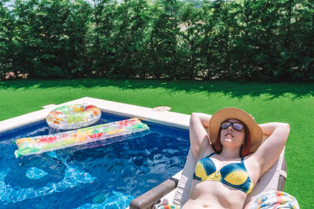 happy young woman in swimming costume and sunglasses lying in a hammock sunbathing by the pool. girl...