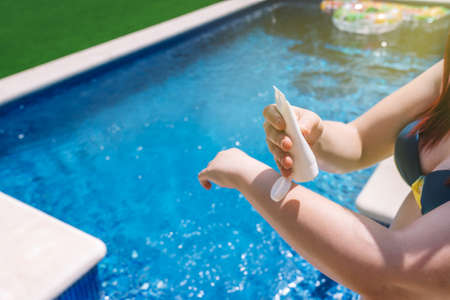 unrecognizable young woman applying sunscreen to the skin on her arms, next to a swimming pool....