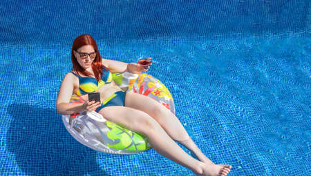 young red-haired girl, looking at her smartphone floating on a ring float inside the pool, while...