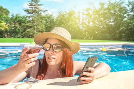 young woman relaxing inside the pool, looking at her mobile phone. girl on holiday sharing the...