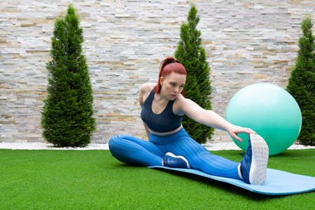 young woman with red hair, practising leg stretching exercises on a mat in the garden of her house....