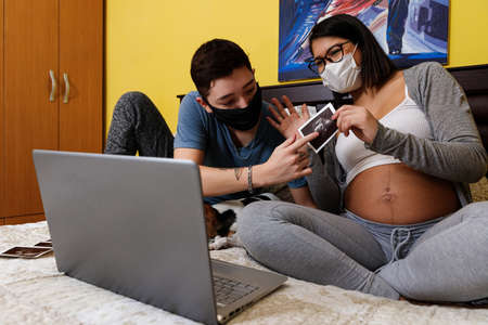 couple of future young parents showing ultrasound by laptop in an online video call, due to the...