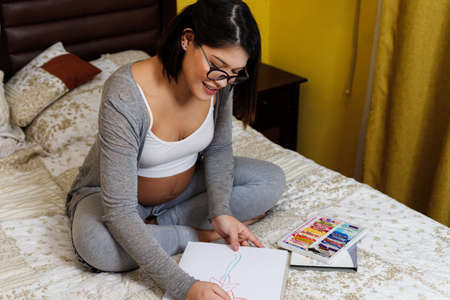 young pregnant mother painting on the bed at home with pastel pencils during quarantine and social distancing by covid-19, happy future mother waiting for her baby.の写真素材