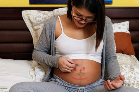 young pregnant mother painting on the bed her abdomen at home with pastel pencils during quarantine...