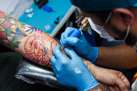 tattooist with his machine tattooing on the arm with the design drawn and using protective mask against covid 19, concept of art and designの写真素材