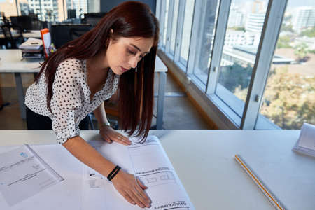young, beautiful and empowered entrepreneurial woman architect, working in the office on her architectural drawings and plans, business concept, design and womanの写真素材