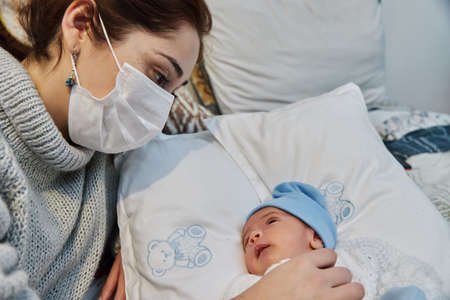 young redheaded mother with preventive health mask and lgbt bracelet, next to her newborn baby playing and laughing together on the bed, concept of family and newbornの写真素材
