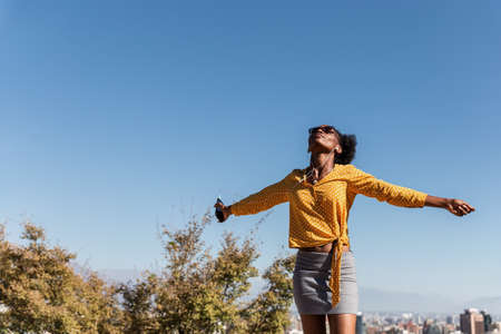 portrait of beautiful young black woman with yellow shirt and sunglasses observing nature in a park, concept of nature, social distancing and portraitの写真素材