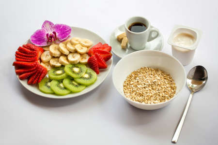 Breakfast of oatmeal, a plate of fruit decorated with a flower orchid, yogurt and coffee on a white backgroundの写真素材