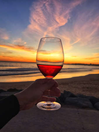 Woman man hand with a glass of red wine on a background of sunset sky and waves on the beach by the ocean in Portugalの写真素材